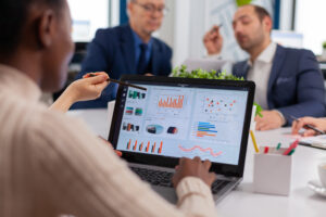 A Black woman points to a laptop screen displaying various business and financial data visualizations, including bar charts, pie charts, and line graphs, during a meeting with several colleagues in a modern office.